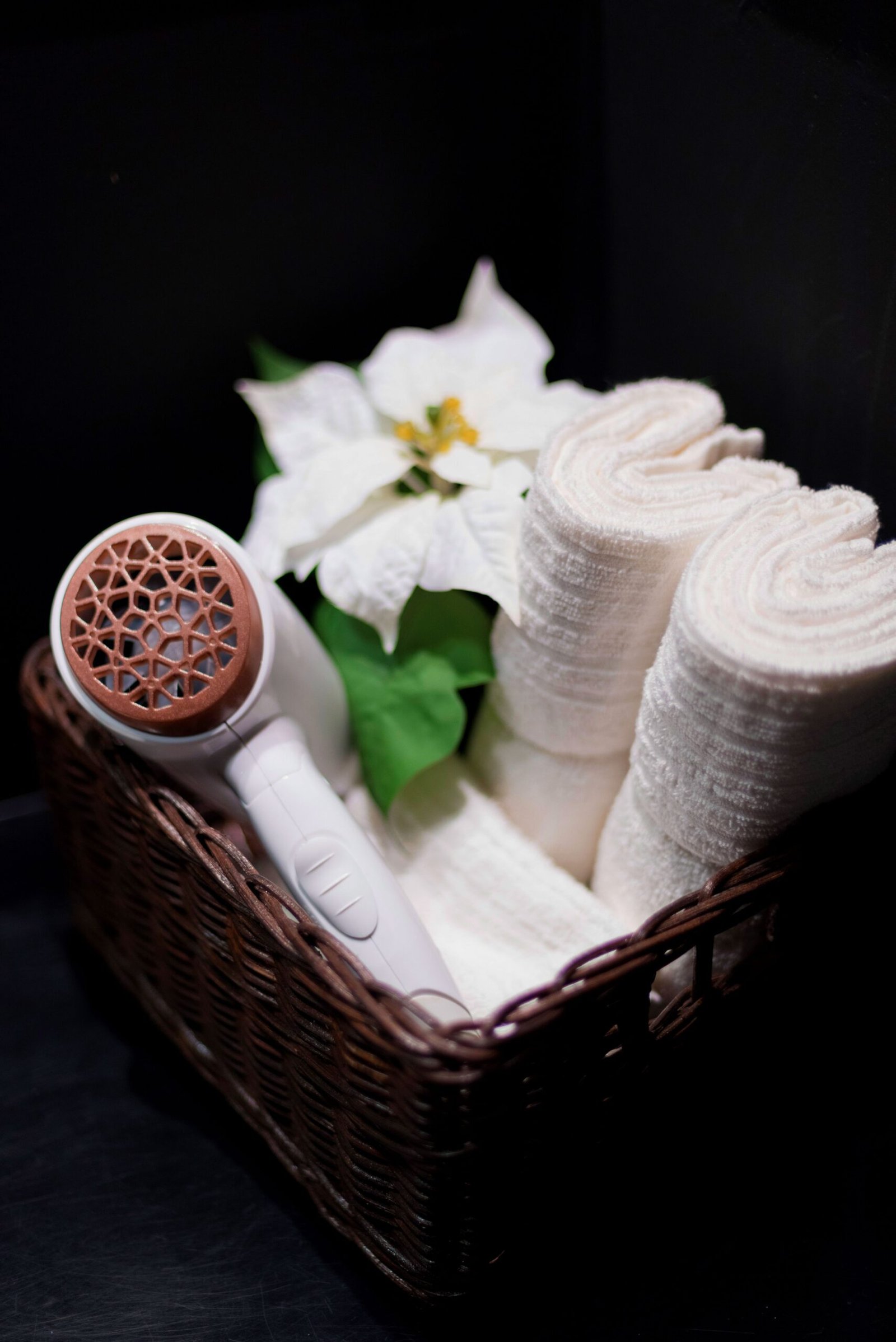 A stylish basket containing rolled towels, a hair dryer, and a decorative flower against a dark background.